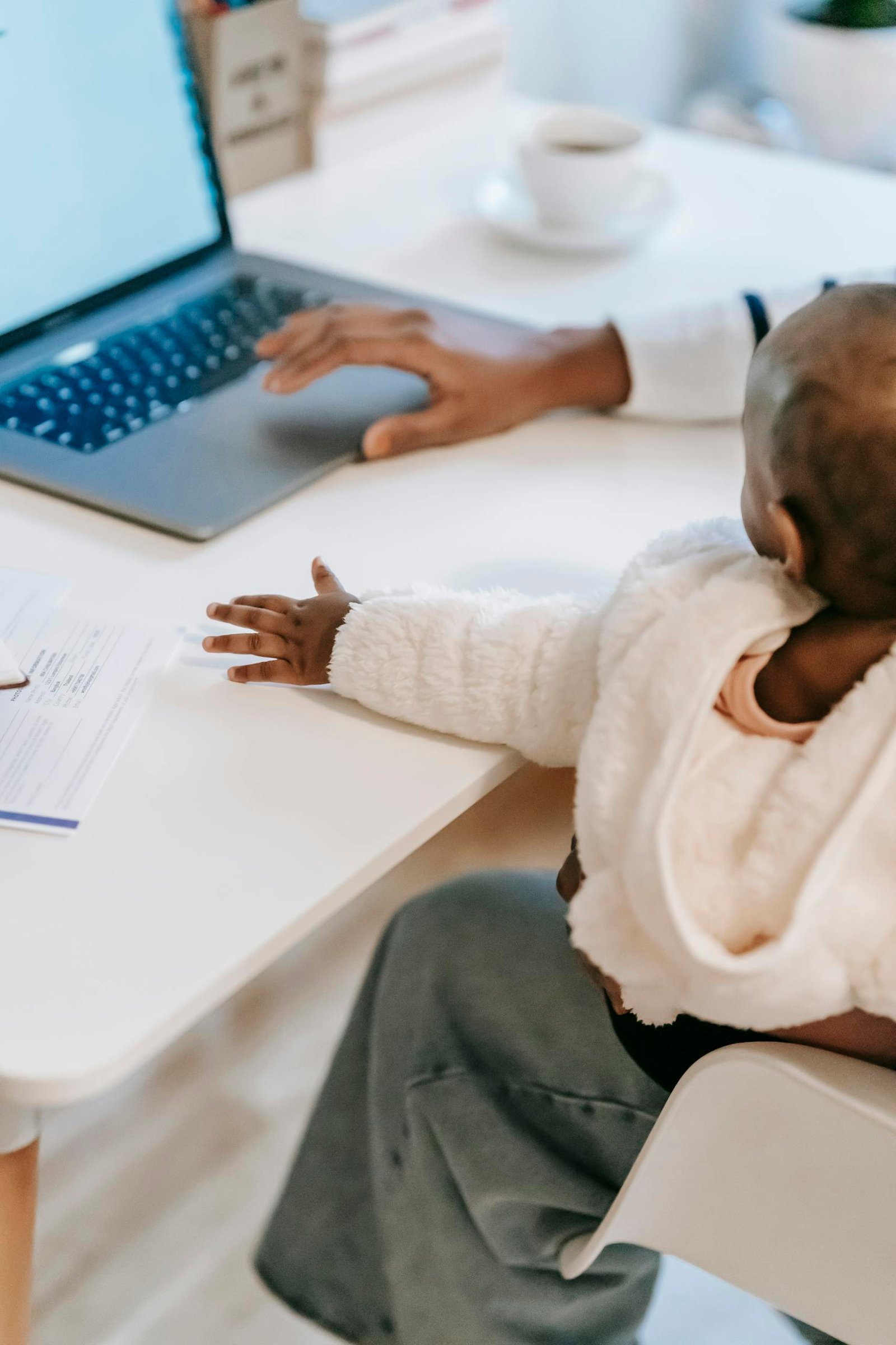 Faceless ethnic female with baby on laps in casual outfit using netbook while sitting in chair near coffee cup and documents at table in light room